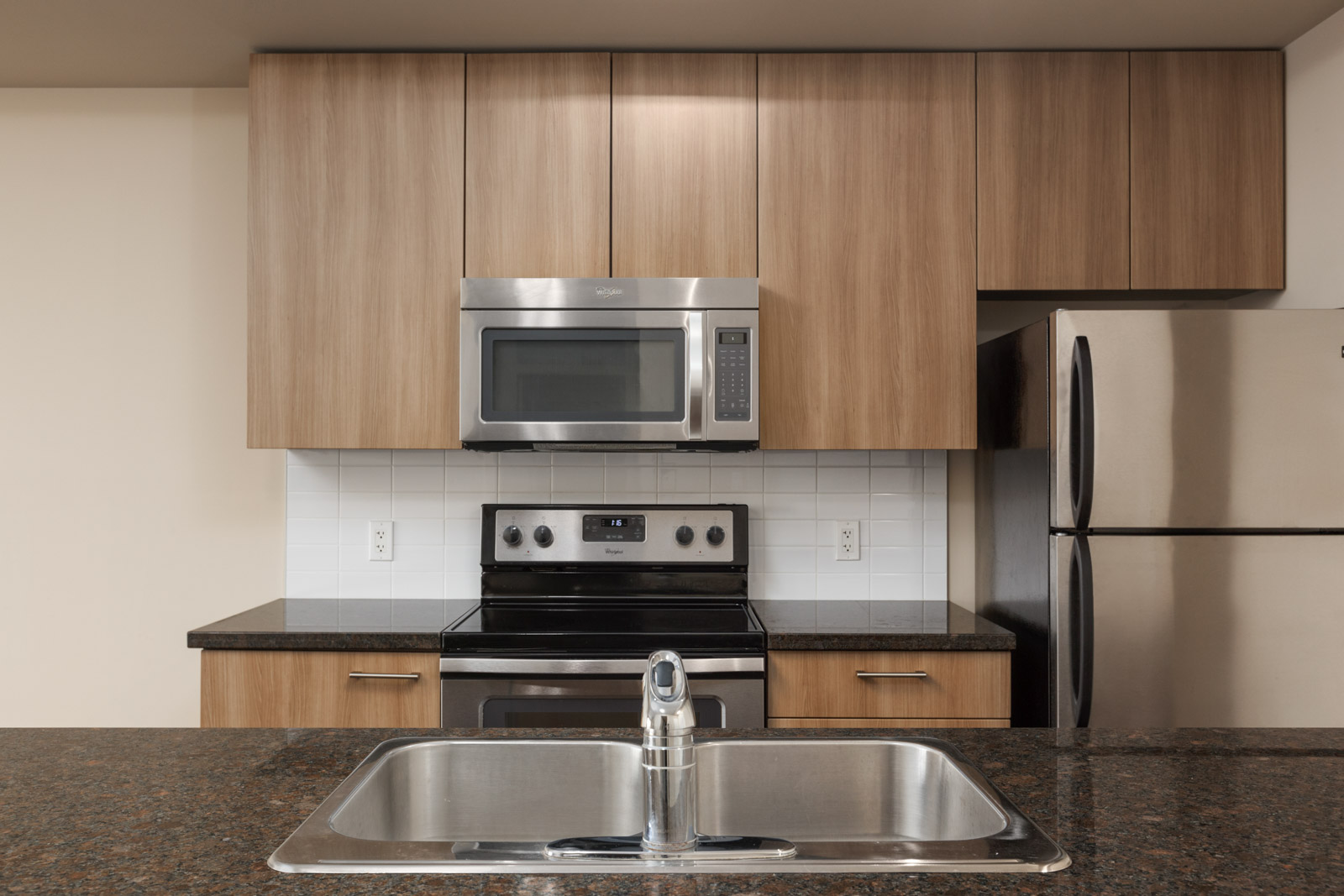 Modern kitchen with wooden cabinets, stainless steel microwave, oven, refrigerator, and a granite countertop with a sink and faucet in the foreground. White tile backsplash.