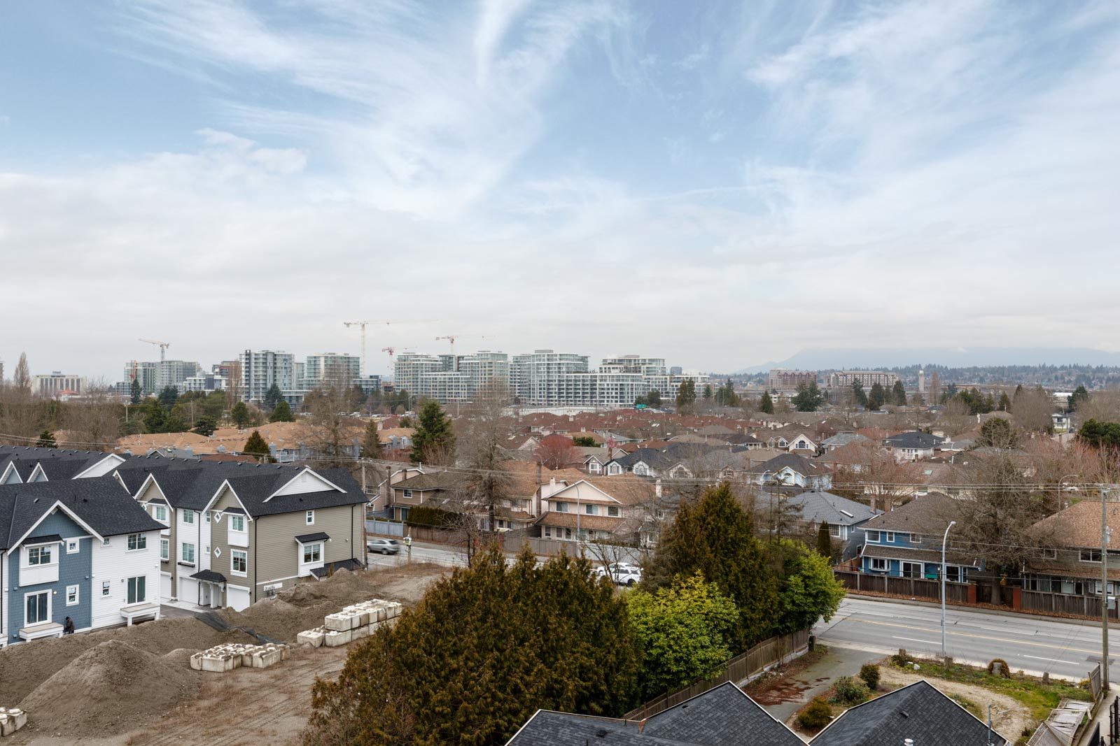 Aerial view of a suburban area with rows of houses, a few cranes in the distance, and a cloudy sky above.