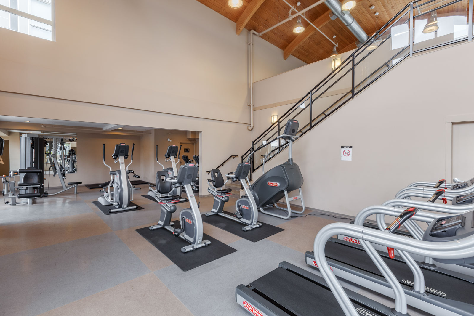 Spacious gym with treadmills, exercise bikes, and weightlifting equipment under a high ceiling with wooden beams.