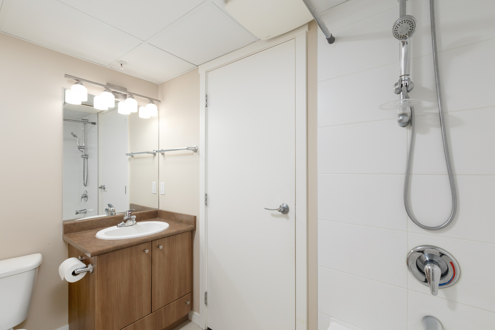A bathroom with a sink, mirror, and wooden cabinet on the left, and a showerhead on the right. The walls are tiled, and a door is in the center.