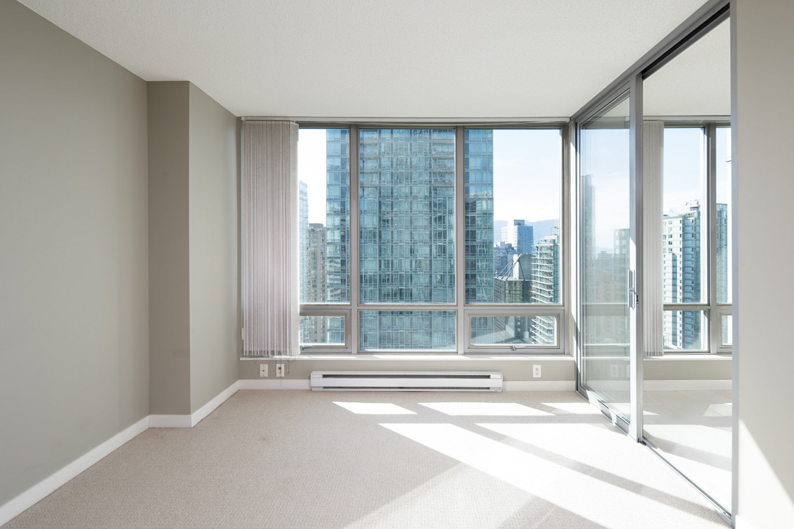 Unfurnished high-rise apartment room with large windows, beige walls, carpet flooring, and a glass door leading to a sunlit balcony with a cityscape view.