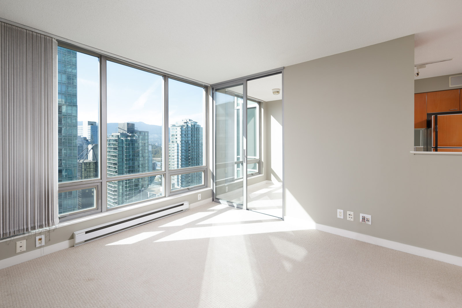 A bright, empty corner room in a high-rise apartment with floor-to-ceiling windows, city views, and beige carpet flooring.