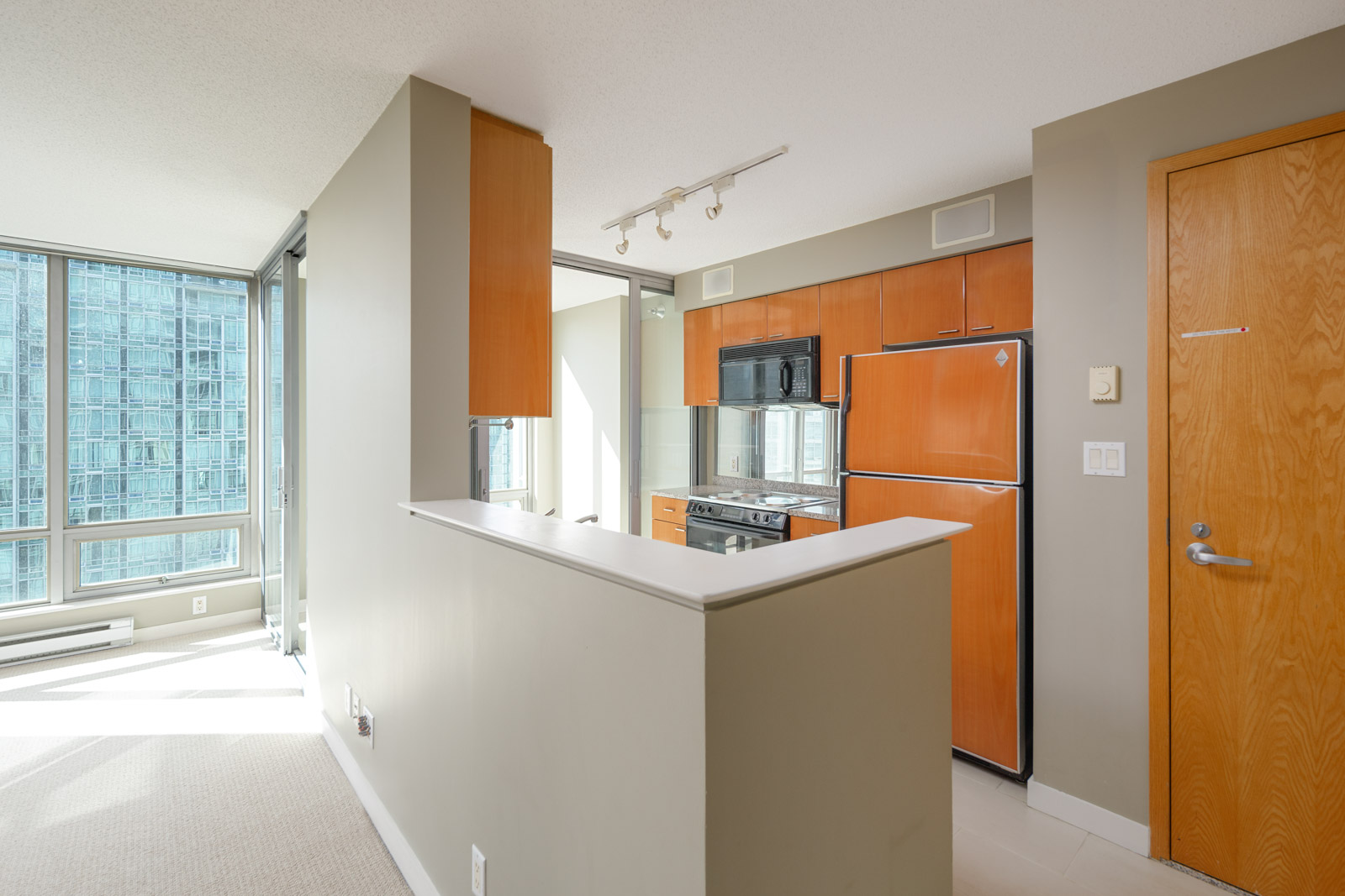 A modern apartment kitchen with light wood cabinets, stainless steel appliances, and a partial wall dividing it from the living area. Large windows let in natural light.