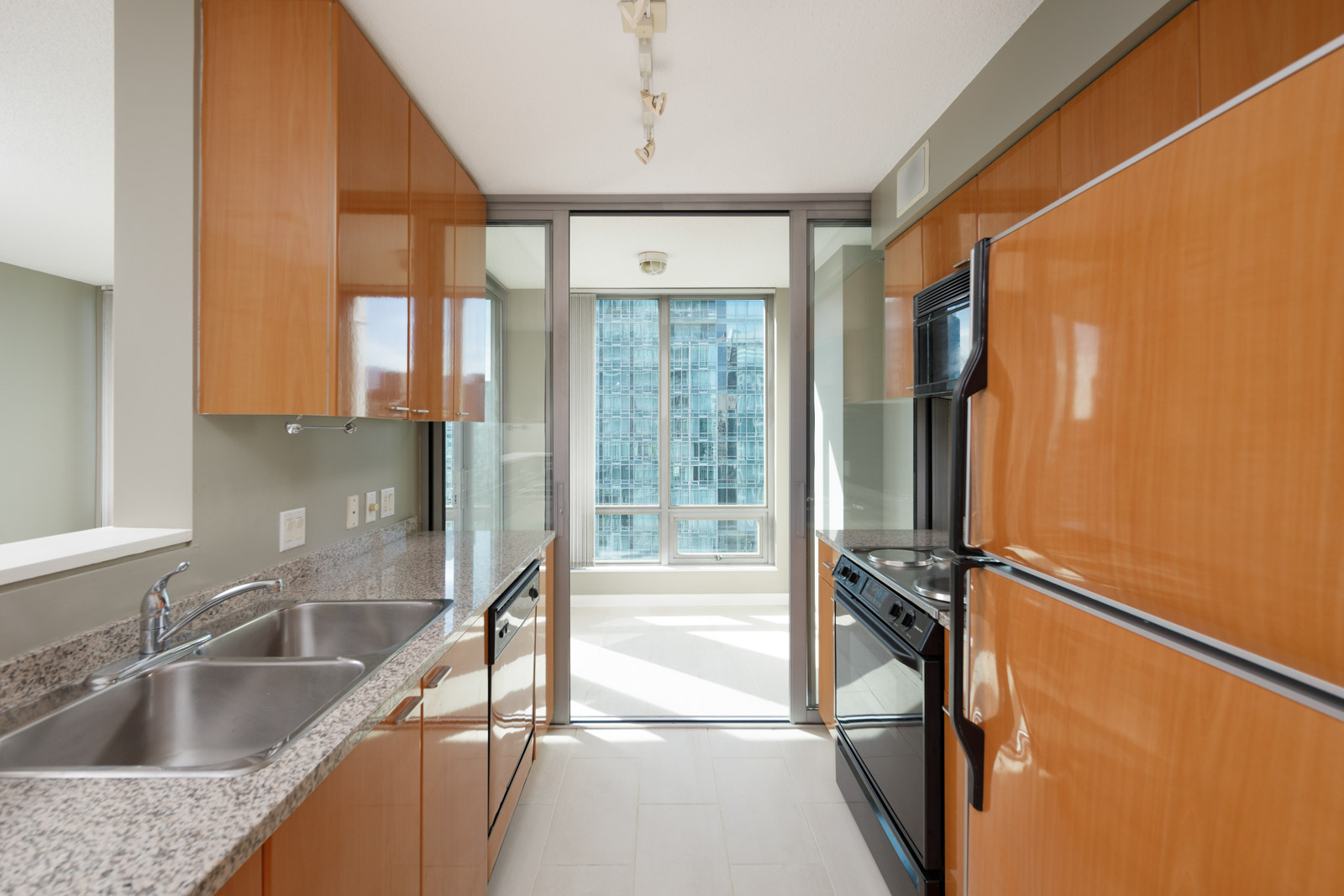 Modern galley kitchen with light wood cabinets, granite countertops, stainless steel sink, and black appliances, leading to a bright dining area with large windows.