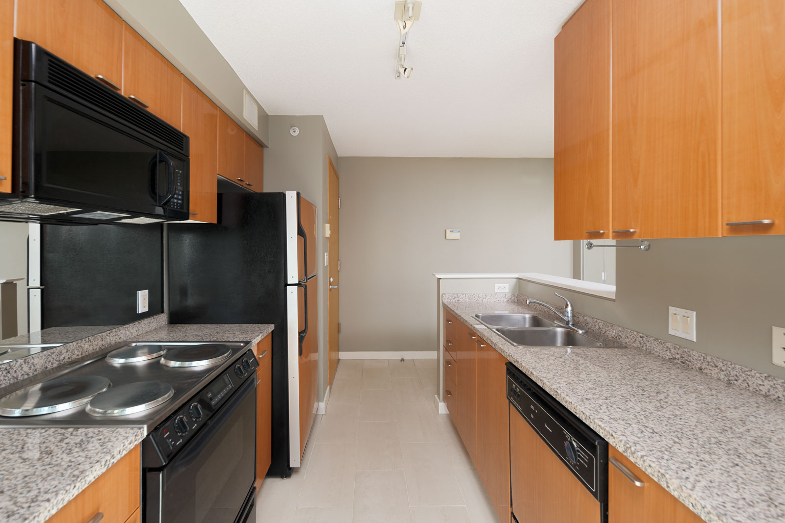 Modern galley kitchen with wood cabinets, granite countertops, black appliances, stainless steel sink, and neutral walls.