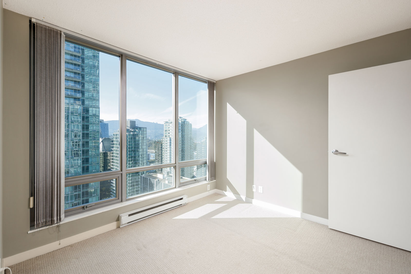 Empty room with beige carpet, large floor-to-ceiling windows, city skyline view, open white door, and neutral-colored walls. Sunlight streaming in through windows.