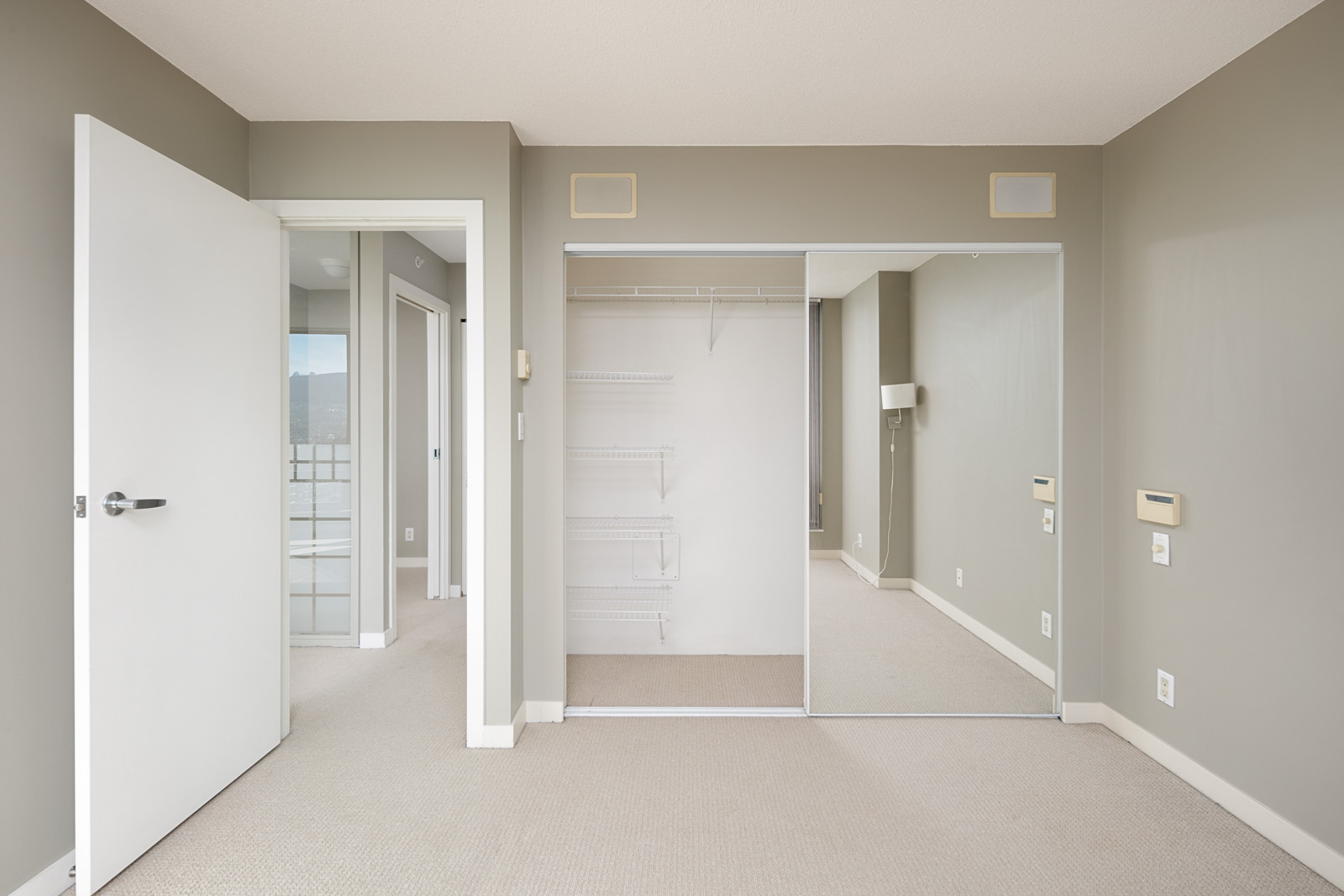 Empty bedroom with beige carpet, light gray walls, an open closet with mirrored sliding doors, and an open door leading to another room.