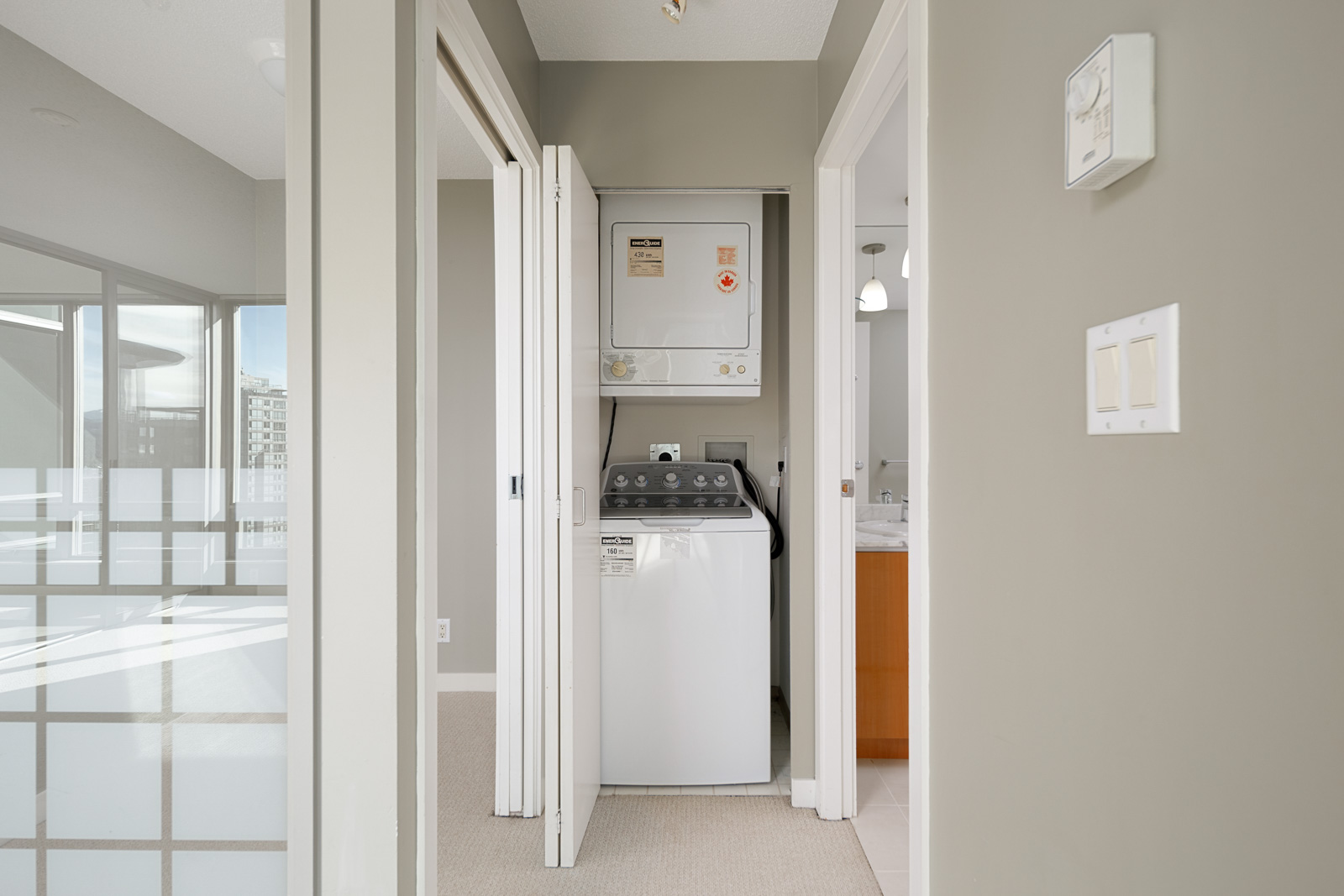 A compact laundry area with a stacked washer and dryer is situated in a hallway between rooms with beige walls and white trim.