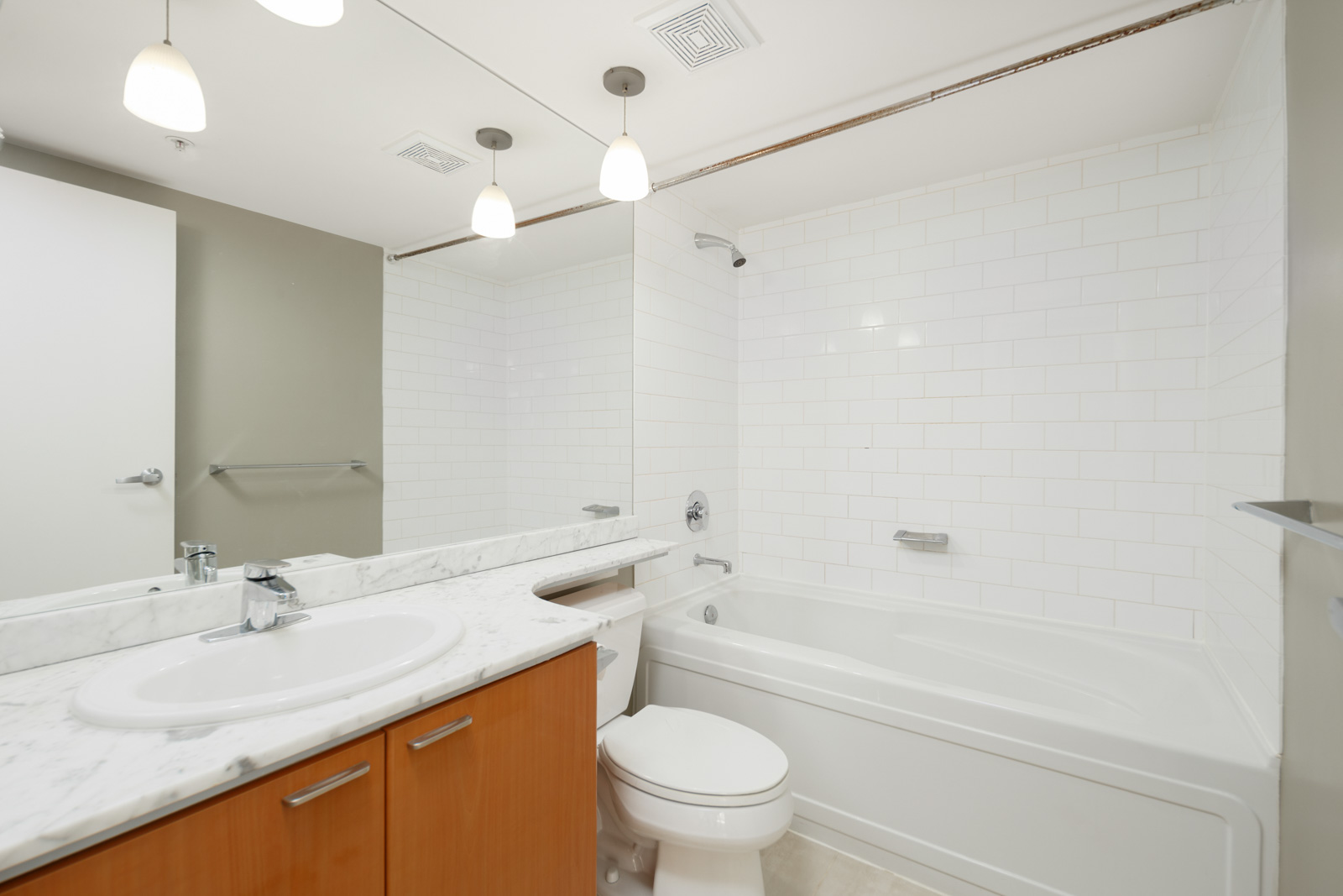 Modern bathroom with white tile walls, a bathtub with shower, a toilet, and a vanity with a sink and marble countertop under a large mirror.