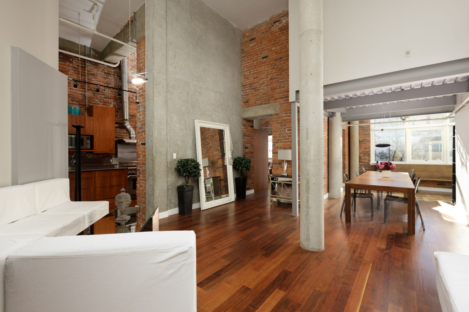 Modern loft apartment with high ceilings, exposed brick walls, and wood flooring. The room features a kitchen area, dining table, white sofas, large mirror, and potted plants.