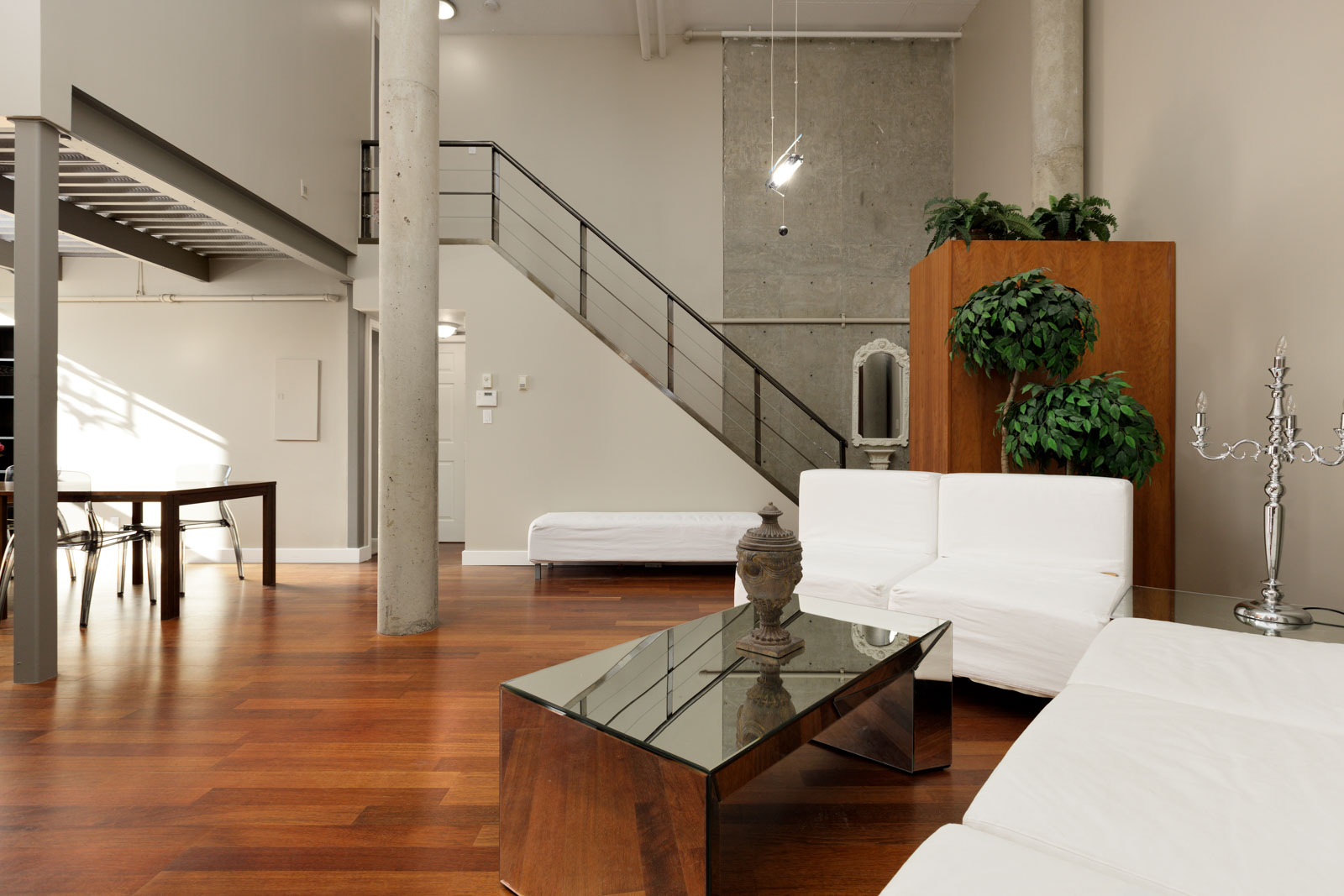 Modern living room with white seating, a glass coffee table, wooden flooring, and minimalist decor. Stairs and a loft area are visible in the background.