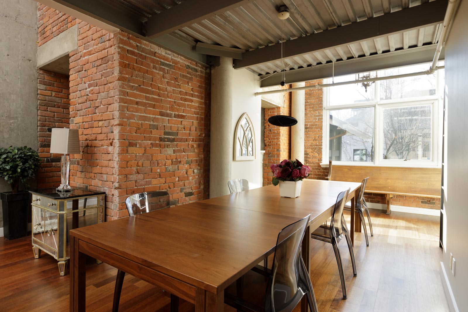 A dining area with a wooden table, clear chairs, a brick wall, and a large window. A potted plant and a side table with a lamp are in the background.