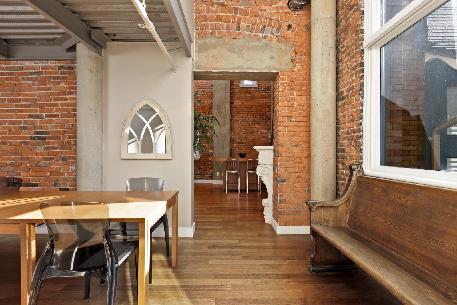 A modern interior with exposed brick walls, wooden floors, and a mix of contemporary and antique furniture. A wooden table with chairs is in the foreground, and a decorative bench is on the right.