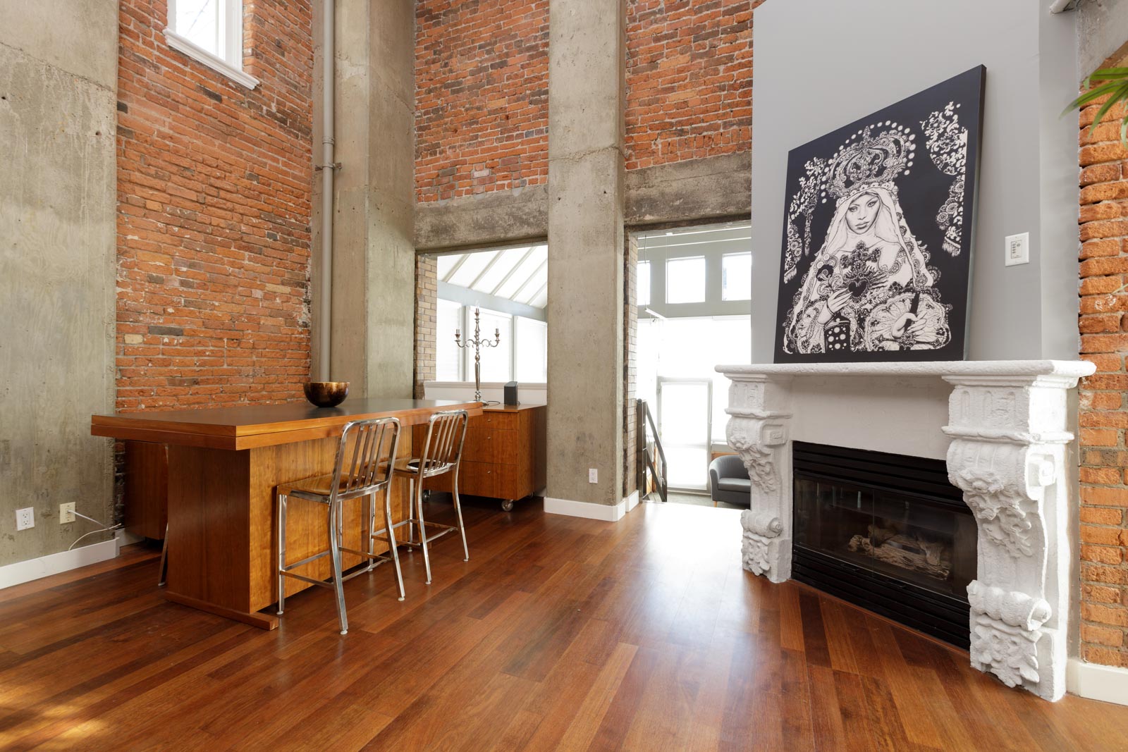 A modern dining area with brick walls features a wooden table, metal chairs, a white fireplace with a black and white artwork above it, and a large window allowing natural light to fill the space.