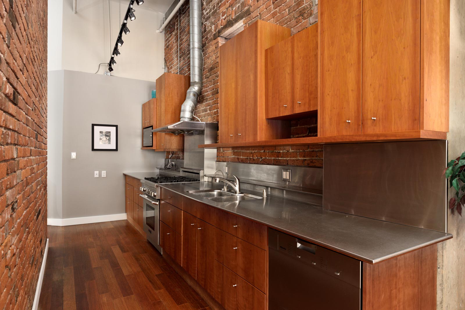 A modern kitchen with wooden cabinetry, stainless steel countertops and appliances, brick walls, a hardwood floor, and a framed picture on a gray wall.