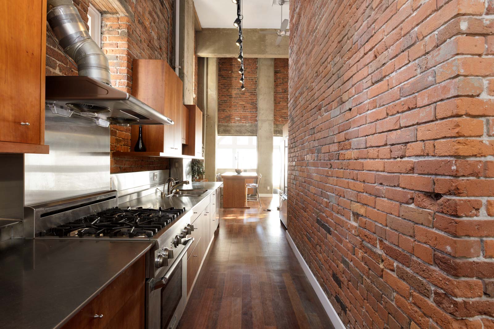 A narrow kitchen with exposed brick walls, stainless steel appliances, and wooden cabinets. The space features a gas stove, backsplash, and track lighting, leading to a sunlit area with a dining table.