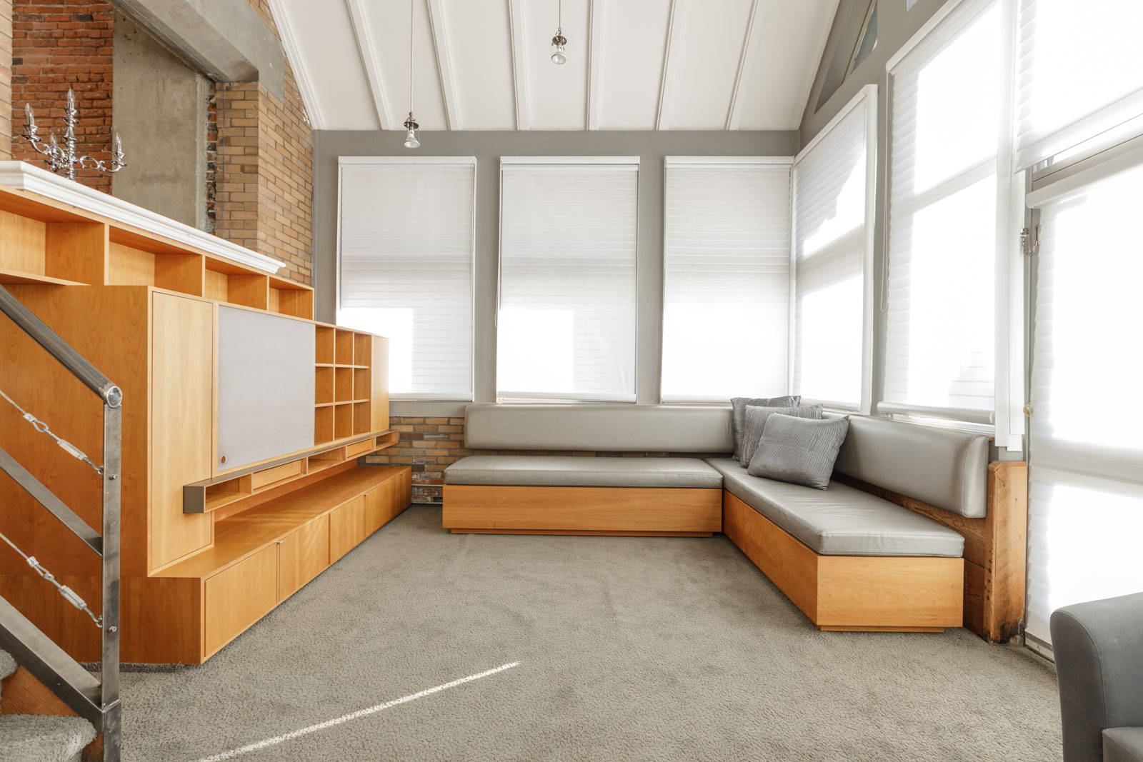 Modern living room with light grey carpet, L-shaped grey sofa, large windows with blinds, wooden storage unit, and staircase with a metal railing.