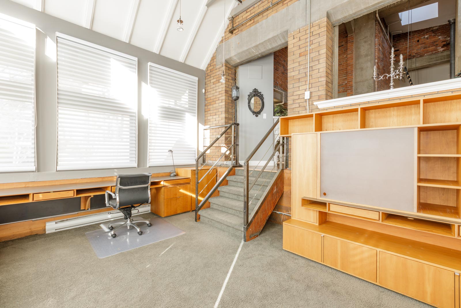 A modern office space with a desk, chair, and built-in wooden shelving. Stairs lead to a second level, and large windows with blinds allow natural light in.