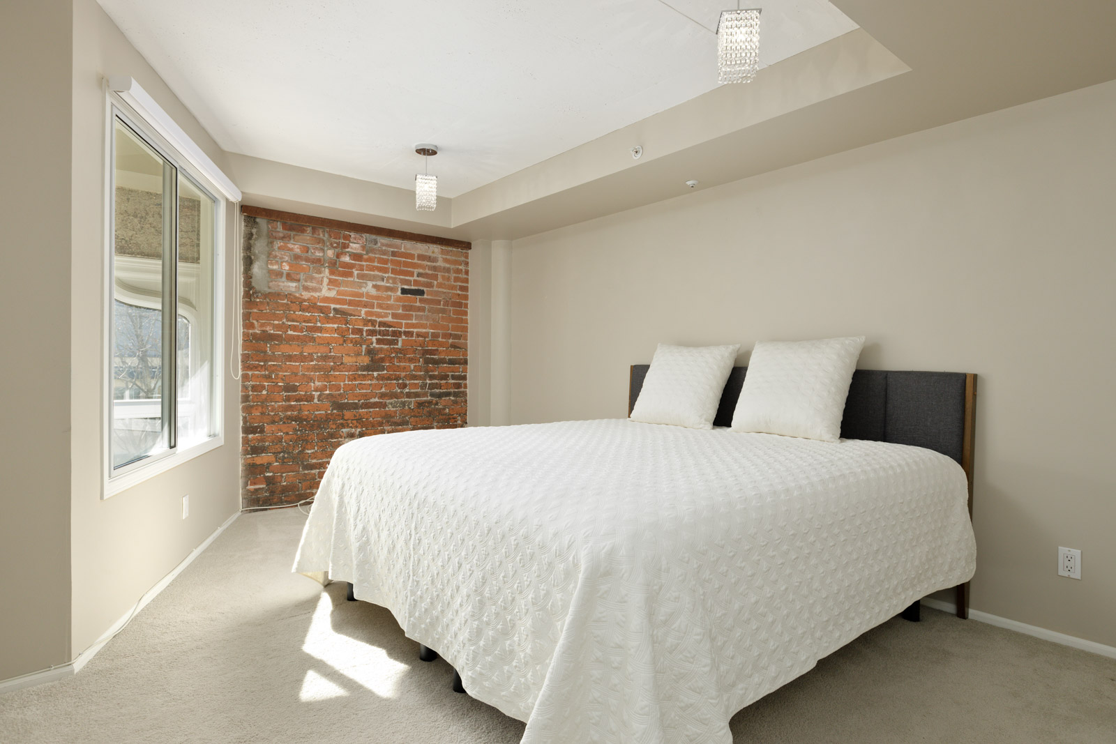 A bedroom with a bed covered in a white quilt, two white pillows, a red brick accent wall, carpeted floor, a large window, and ceiling lights.