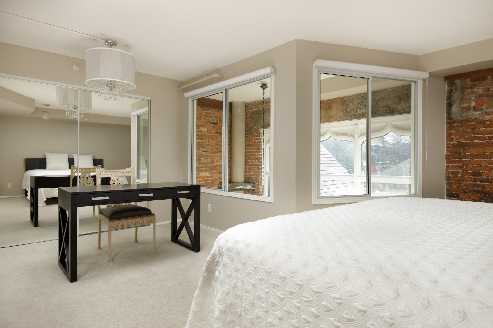 A cozy bedroom with a bed, a black desk with a chair, large windows, mirrored sliding closet doors, and exposed brick accents.