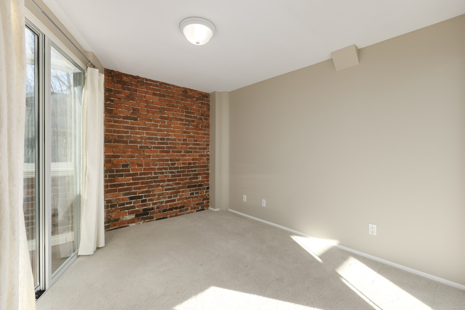 Empty room with beige walls, a brick accent wall, a sliding glass door with white curtains, and beige carpet. The room is lit by a ceiling light and natural light from the window.