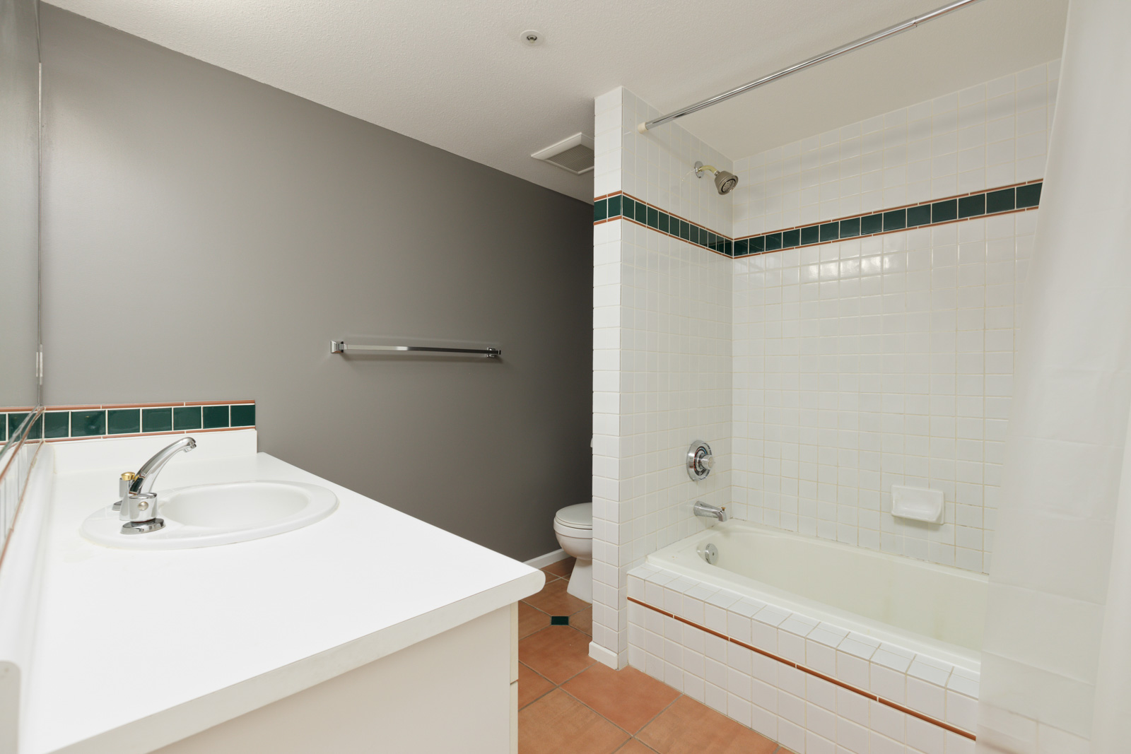 A bathroom with a white sink and countertop, a towel rack, a toilet, and a bathtub-shower combination with a white tiled wall and green accent tiles.