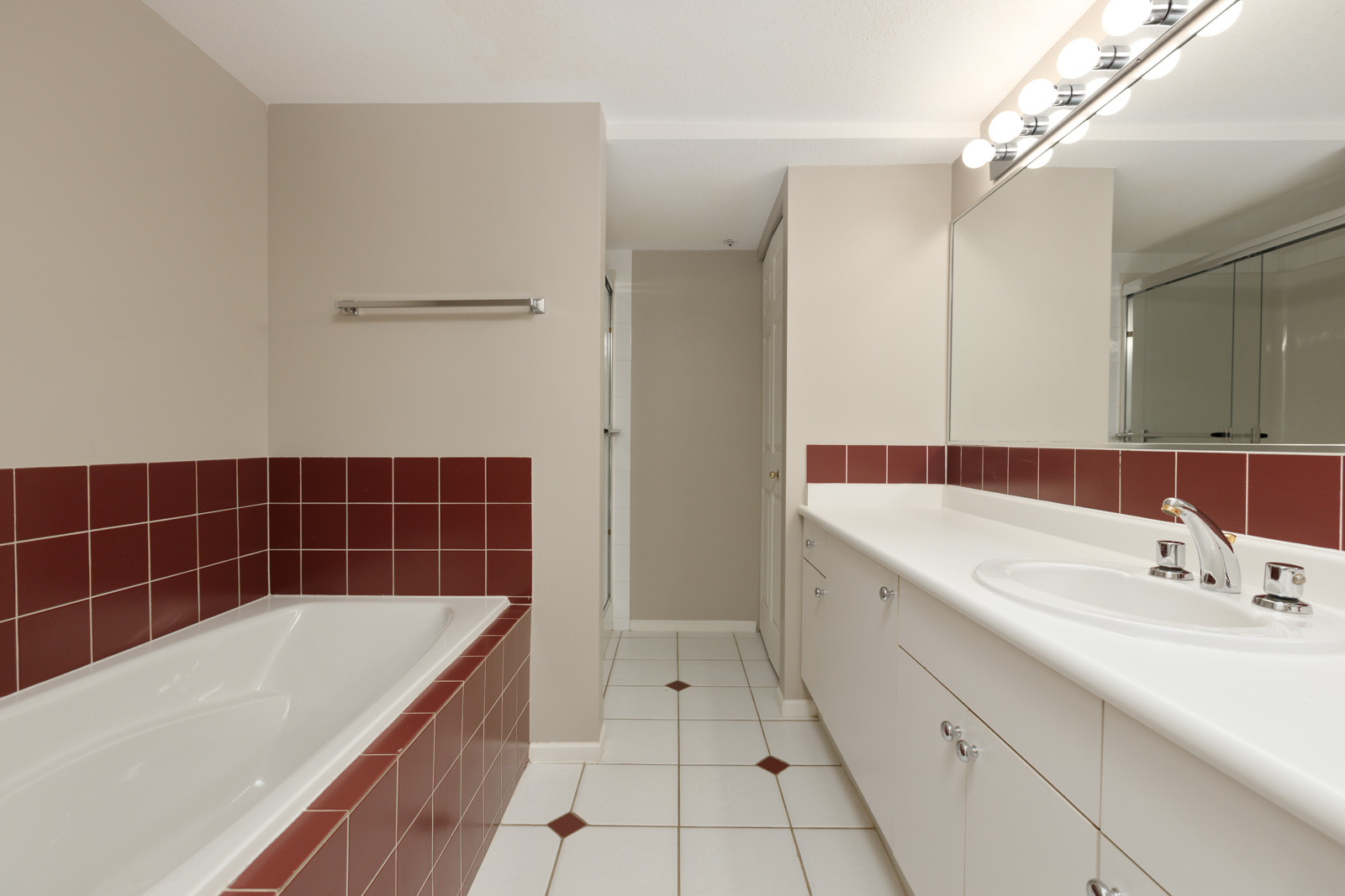 A bathroom with a white double sink vanity, a large mirror, a tub with red tile surround, and beige walls.