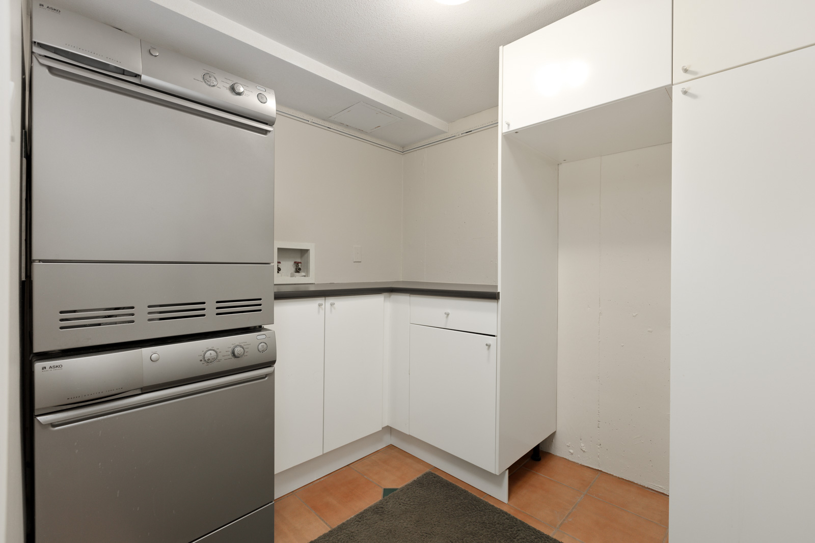 A small laundry room features stainless steel washer and dryer units stacked on the left, white cabinets for storage, a countertop, and tiled flooring.