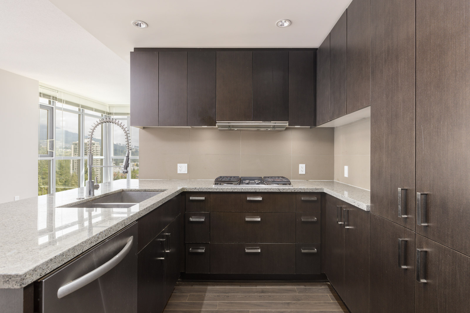 Modern kitchen with dark wood cabinets, silver appliances, and a light granite countertop. Contains a sink, stove, and cabinets with a view of large windows.