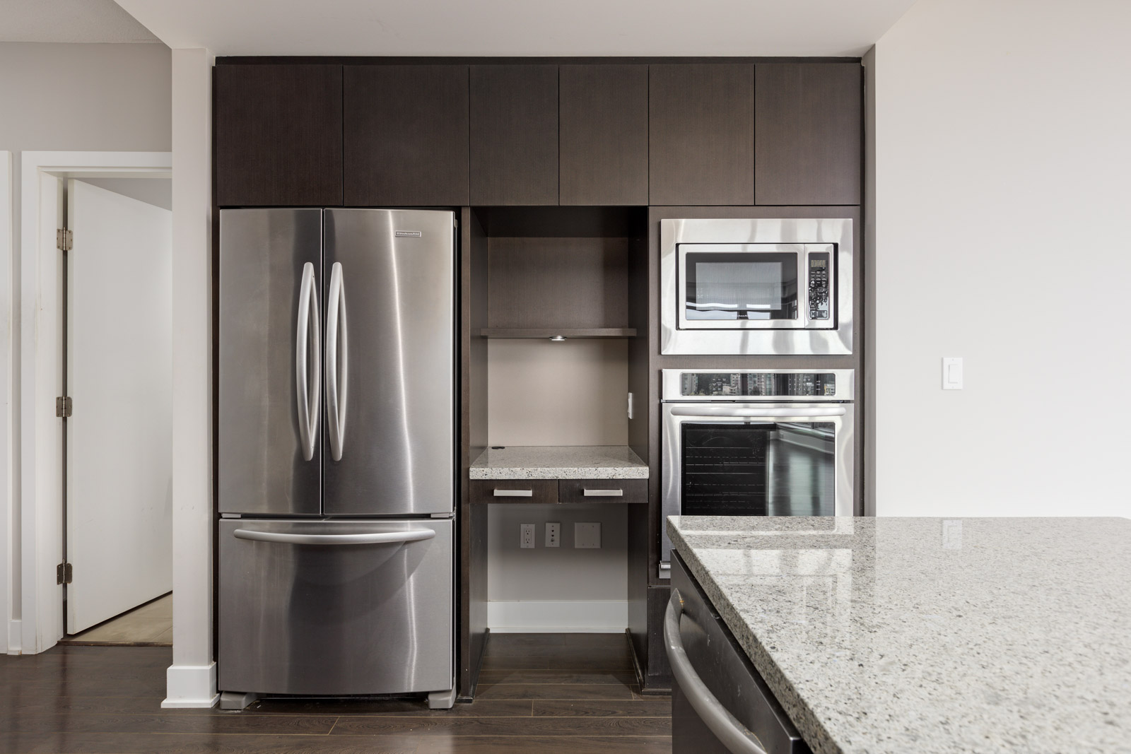 Modern kitchen with stainless steel refrigerator, microwave, and oven built into dark wood cabinets. Gray granite countertops are visible.