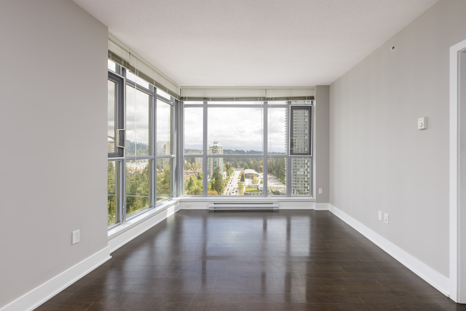 Empty room with large floor-to-ceiling windows, offering a view of a cityscape and distant trees. The room has dark hardwood floors and light gray walls.
