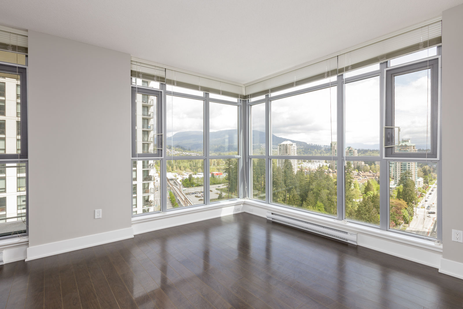 Corner room with large windows offering a view of mountains and city buildings, featuring dark wooden flooring and light gray walls.