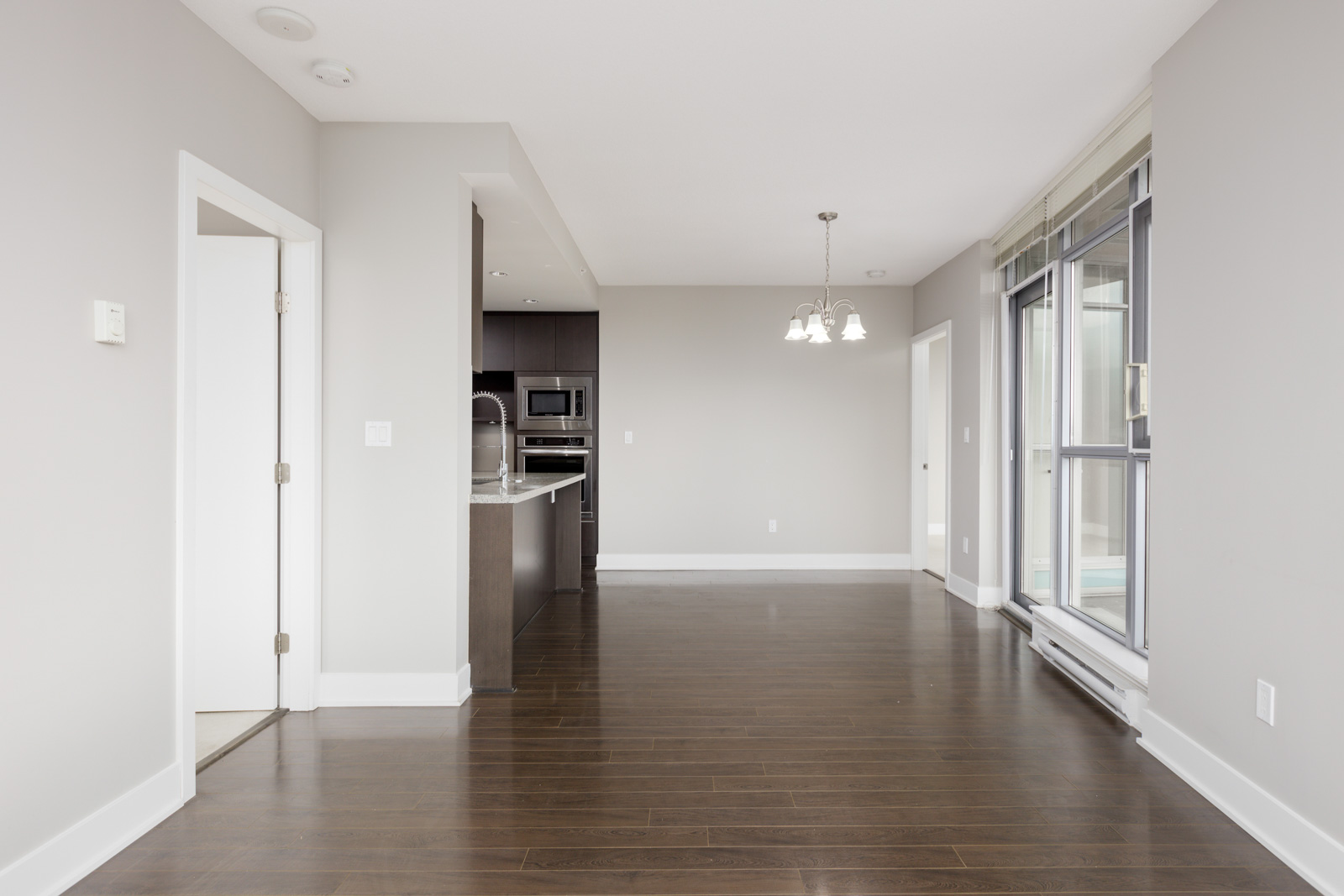 Empty modern apartment interior with hardwood floors, a chandelier, large windows, and an open view into the kitchen area.