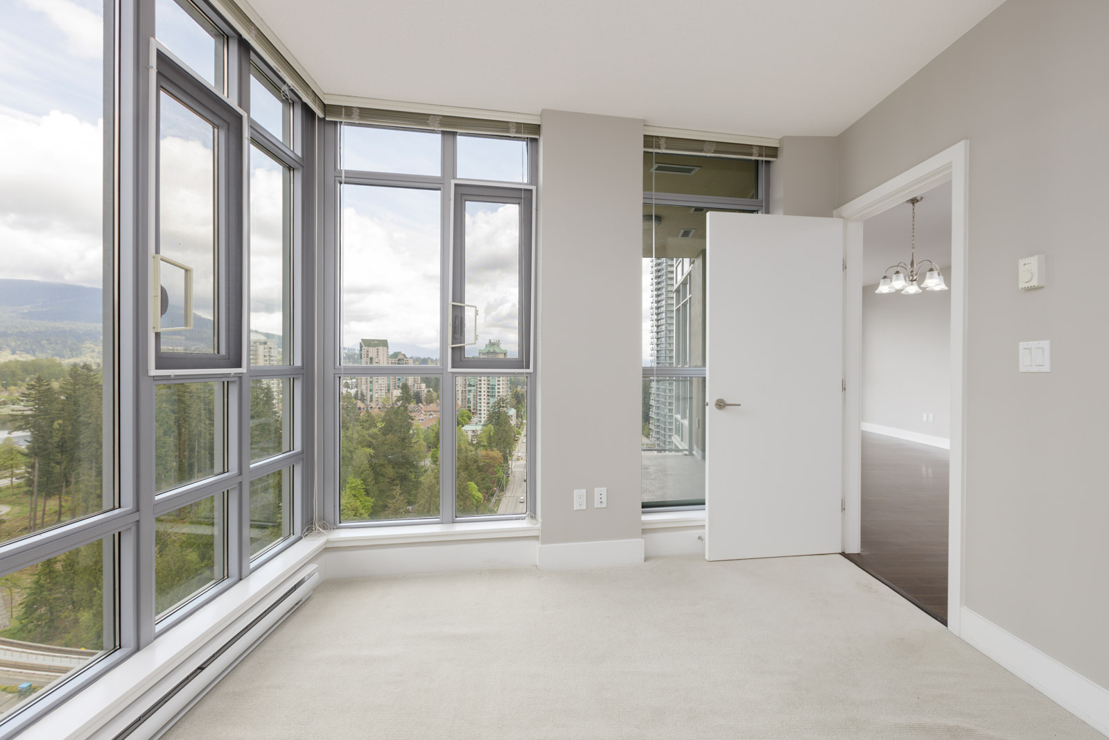 Bright corner room with large windows, a view of trees and buildings, light-colored walls, and an open door leading to a room with dark flooring and a chandelier.