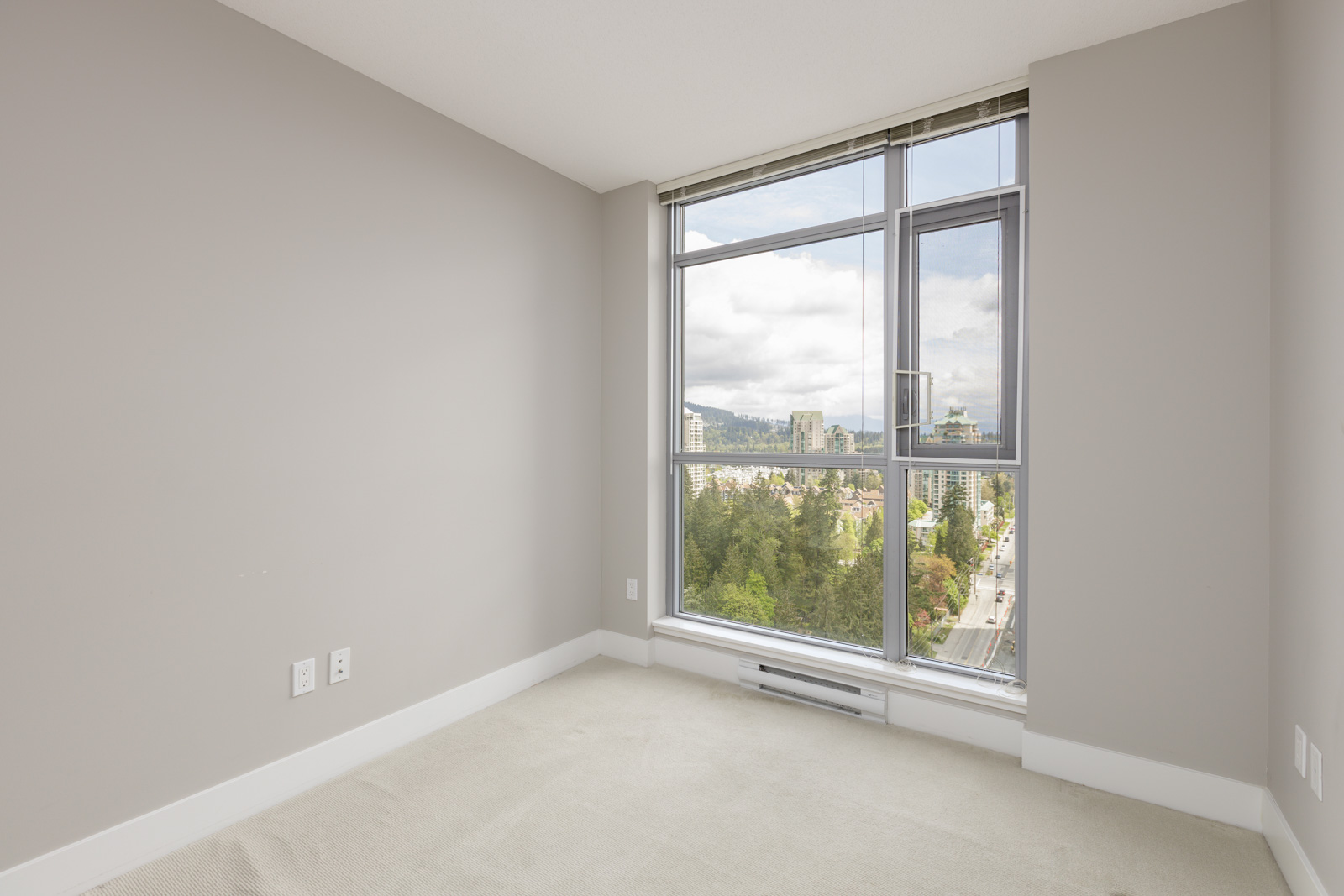 Empty room with gray walls, large window offering a city view, and light carpeted floor. Electrical outlets are visible on the left wall.
