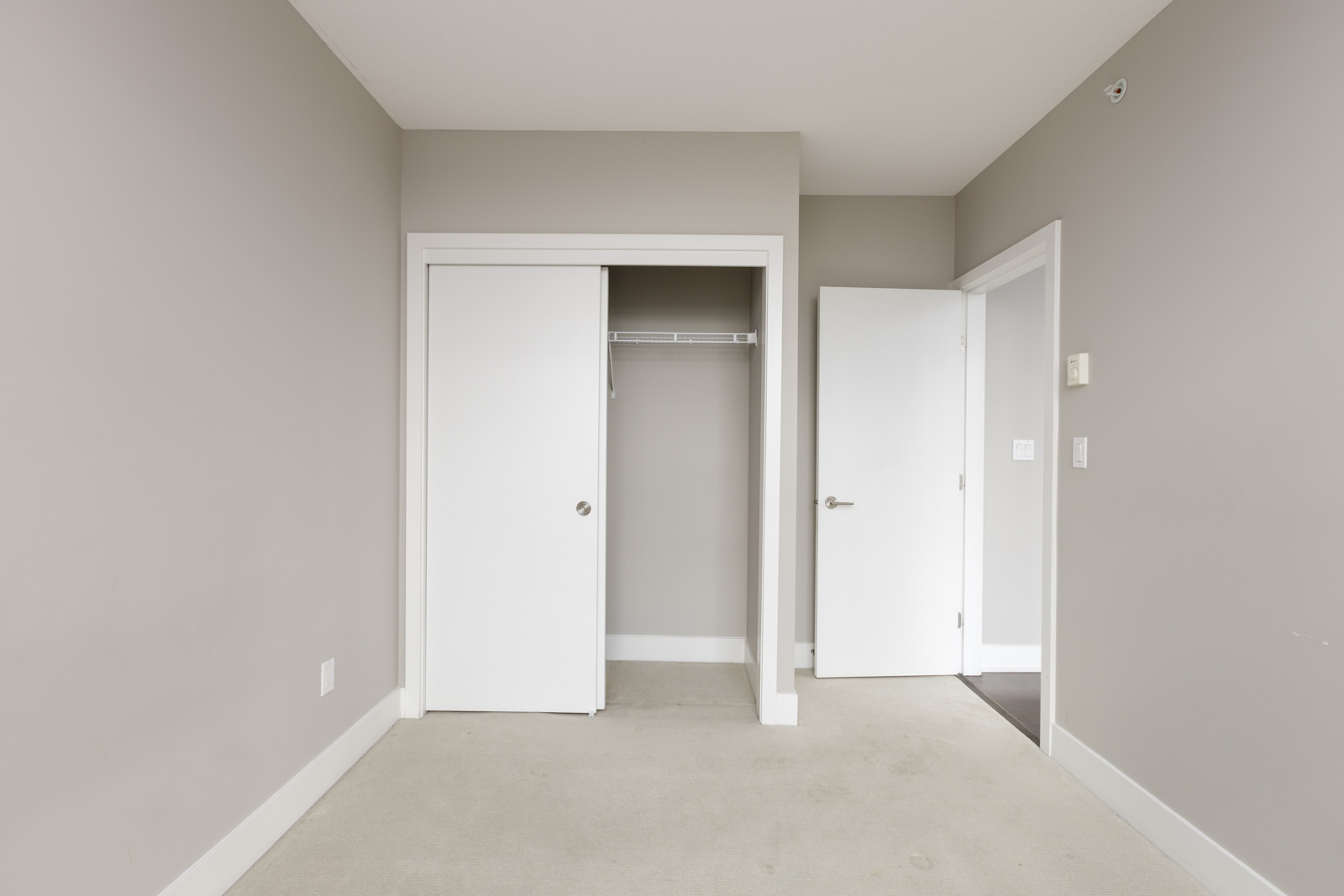 Empty room with beige walls, white trim, and a white closet door. The closet is open, showing a single shelf rack. A white door is ajar leading to another space. Carpeted floor.