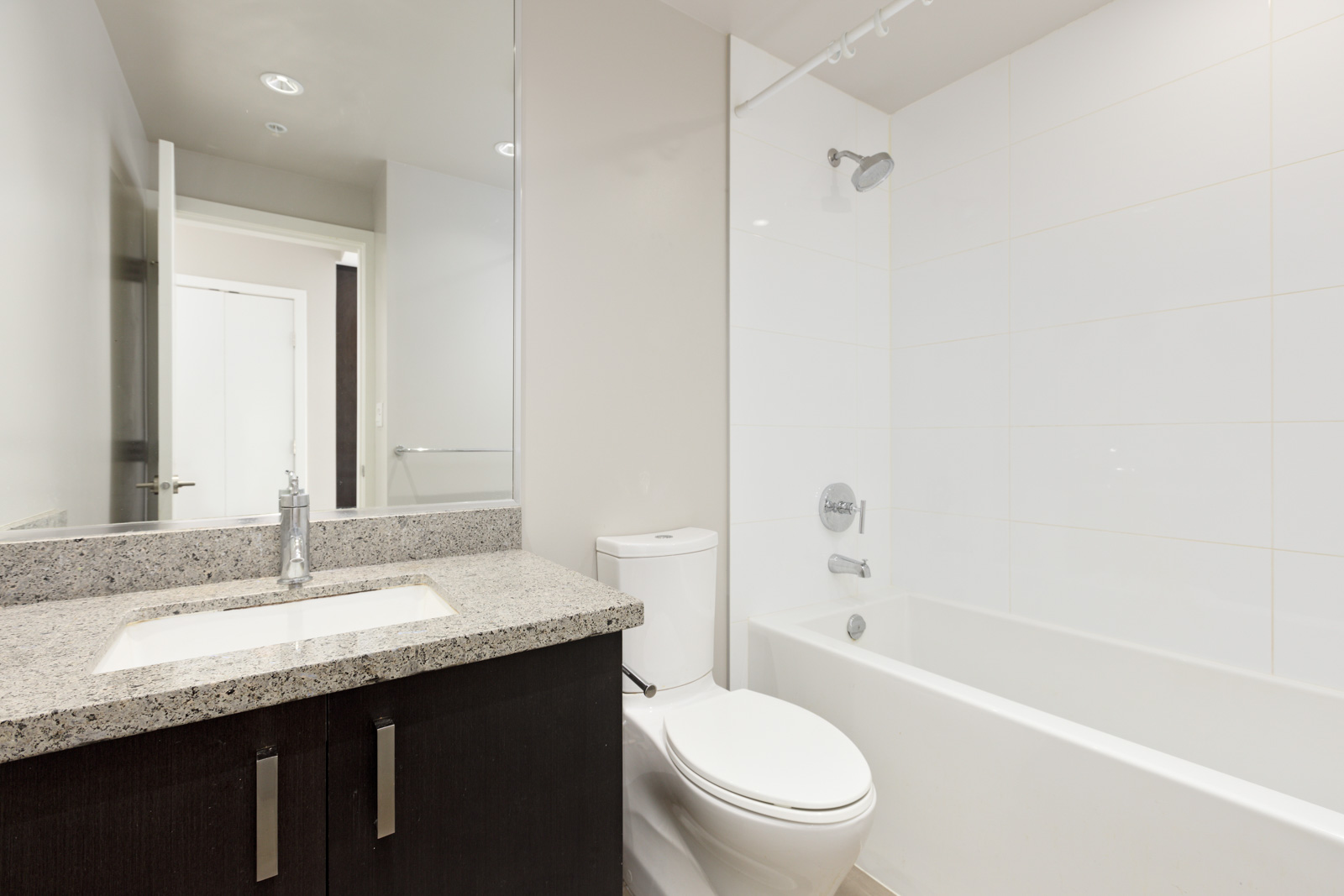 Modern bathroom with a granite countertop, sink, toilet, and a white bathtub with showerhead. Walls and floor are tiled in white. Door visible in the background.