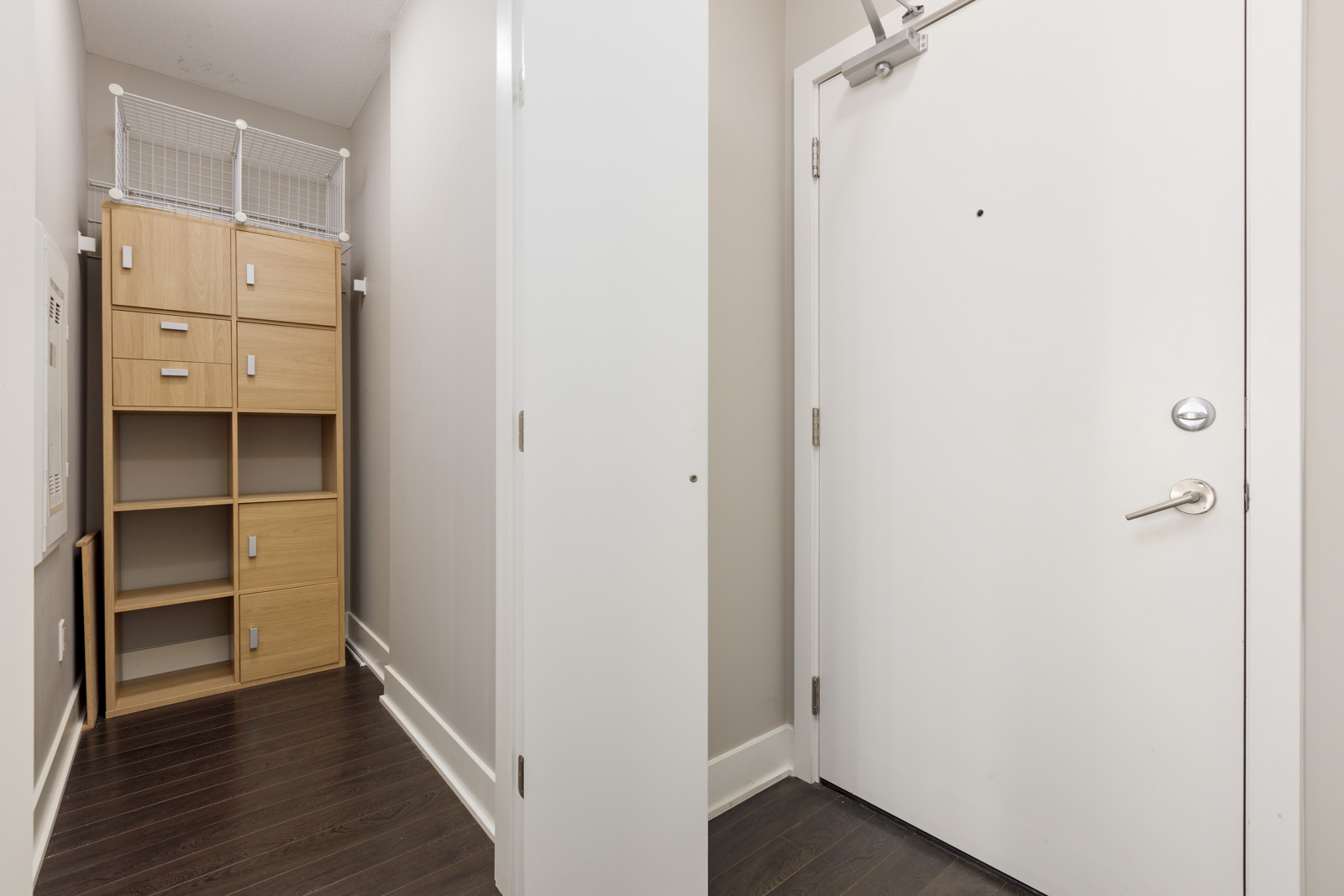 Small hallway with dark wood flooring, a white door on one side, and a light wood storage cabinet with a wire shelf on top.