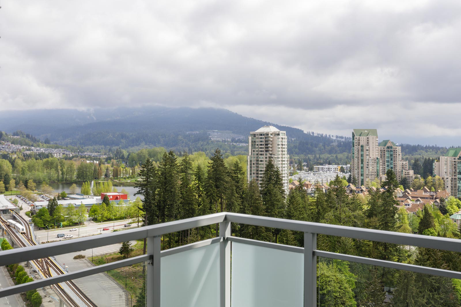 View from a balcony overlooking a cityscape with tall buildings, lush green trees, a railway, and hills under a cloudy sky.