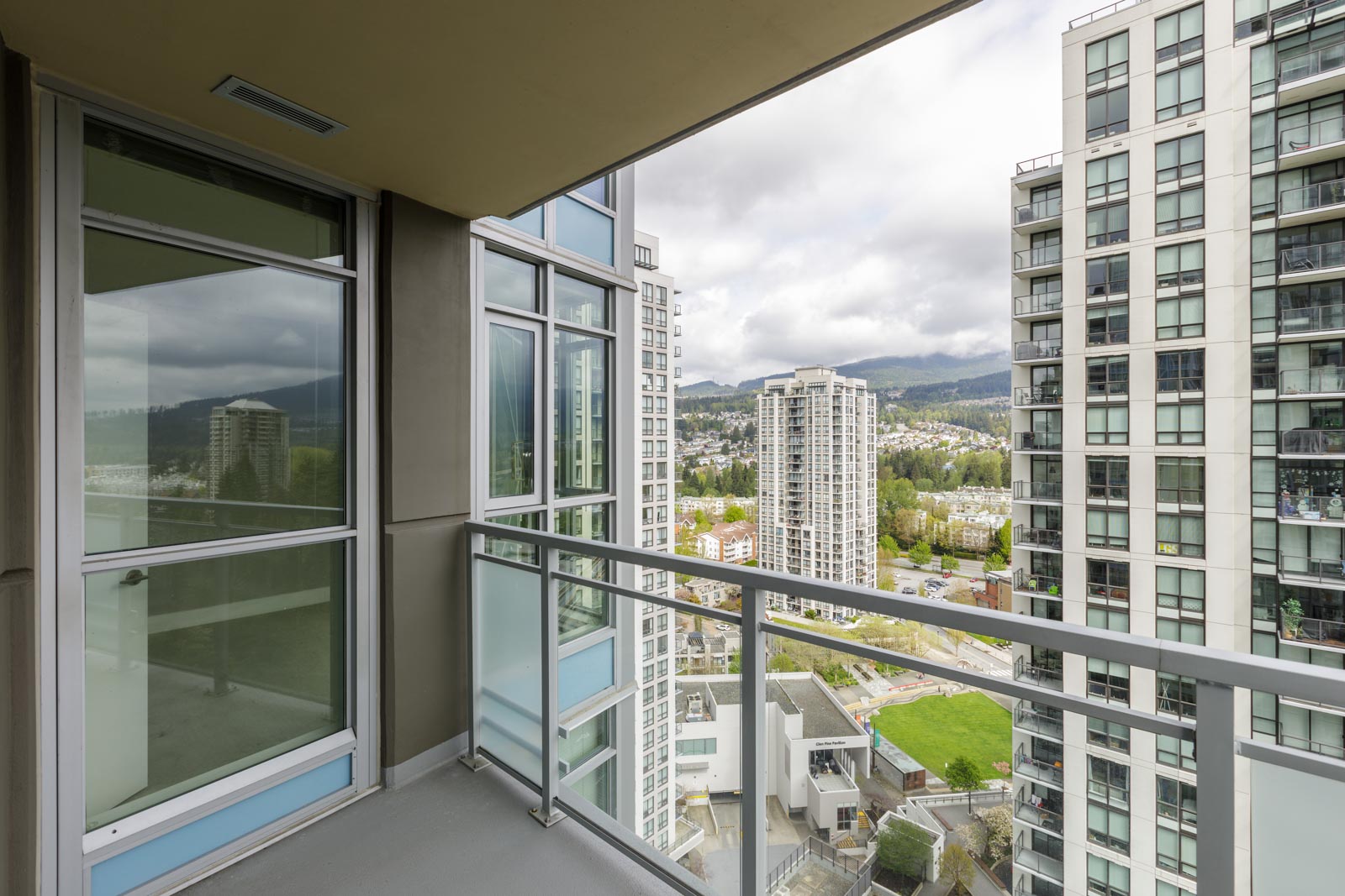 A view from a high-rise balcony showing adjacent tall buildings, a green landscape, and mountains in the background under a cloudy sky.