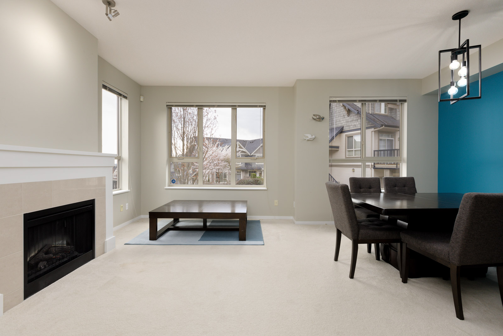 A modern living and dining area with beige carpet, a fireplace, large windows, a coffee table, and a dining table with four chairs. Walls are light-colored with an accent wall in blue.