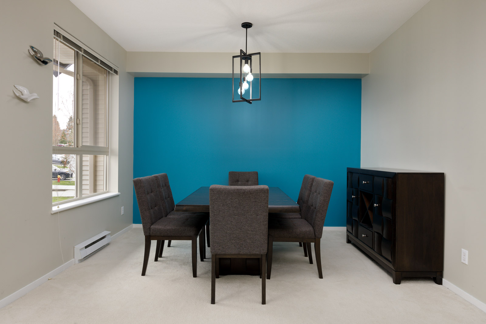 A dining room with a dark wooden table, six grey upholstered chairs, a sideboard, and a modern chandelier against a bold blue accent wall. A window on the left offers natural light.