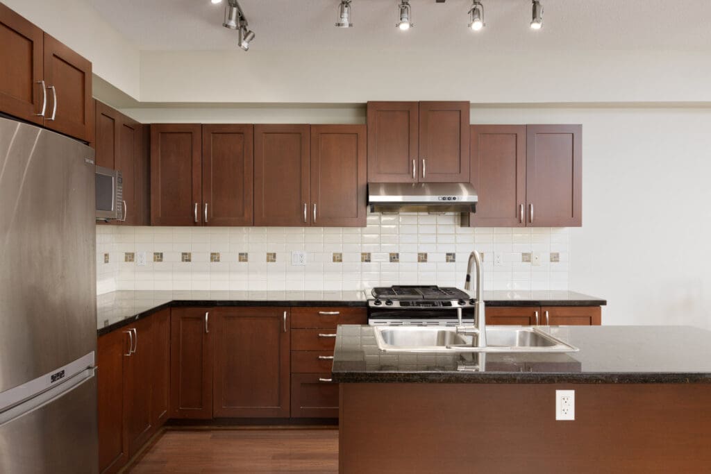 A modern kitchen with dark wood cabinets, stainless steel appliances, a double sink, and a black granite countertop. The backsplash is white tile with decorative accents. Track lighting is installed on the ceiling.
