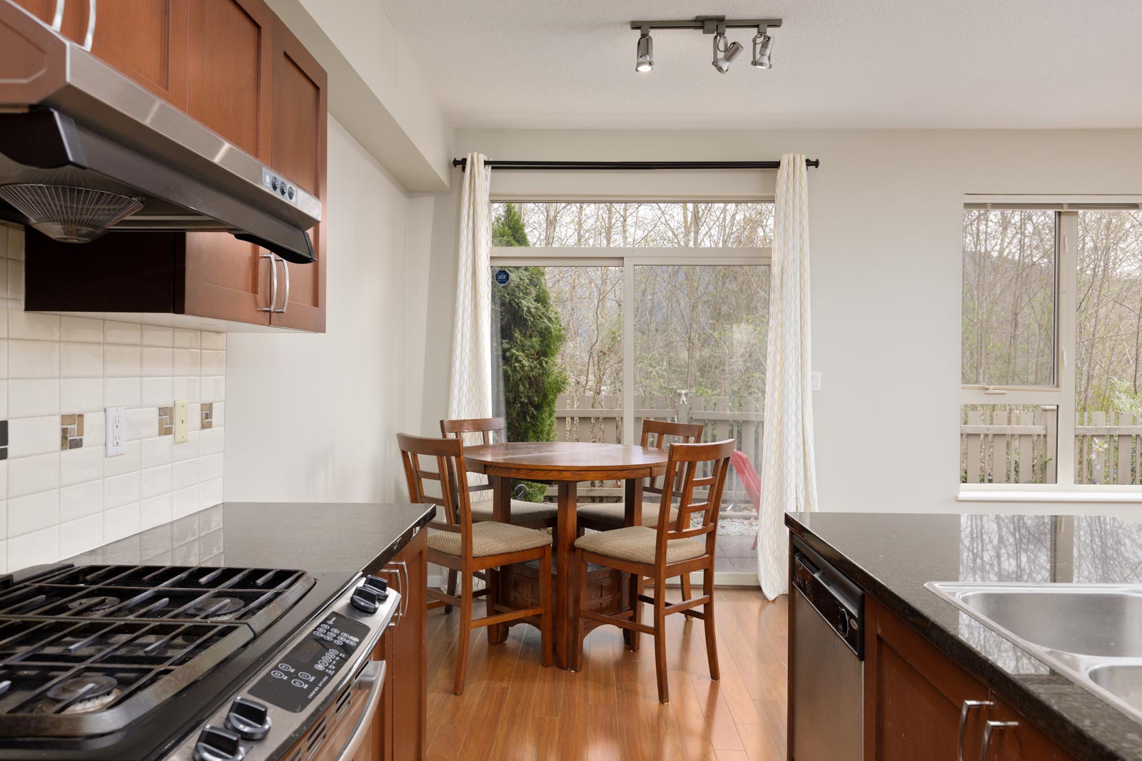 A modern kitchen with wooden cabinets, black countertops, a gas stove, and a round dining table with four chairs near large windows with white curtains.