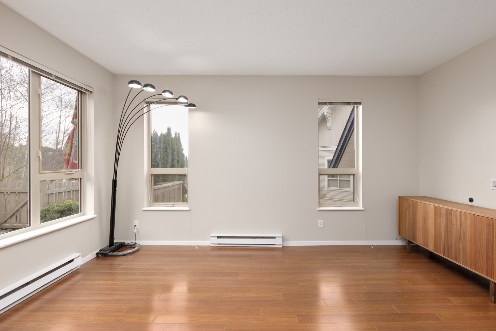 An empty room with wooden floors, a floor lamp, a wooden cabinet, and large windows providing natural light.