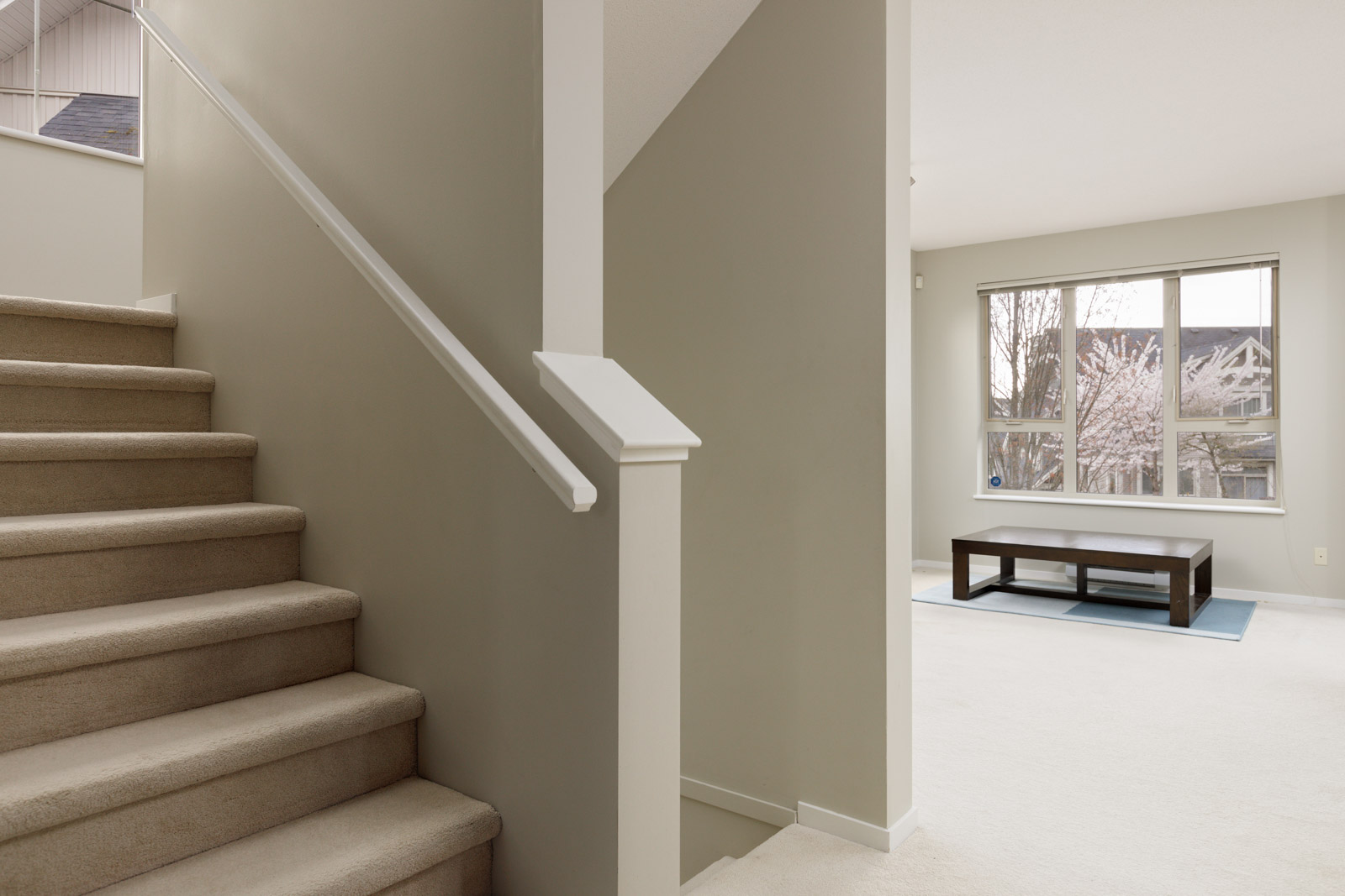 A staircase with beige carpet leads to an upper level. Adjacent is a room with a large window, a coffee table, and a pale carpet.