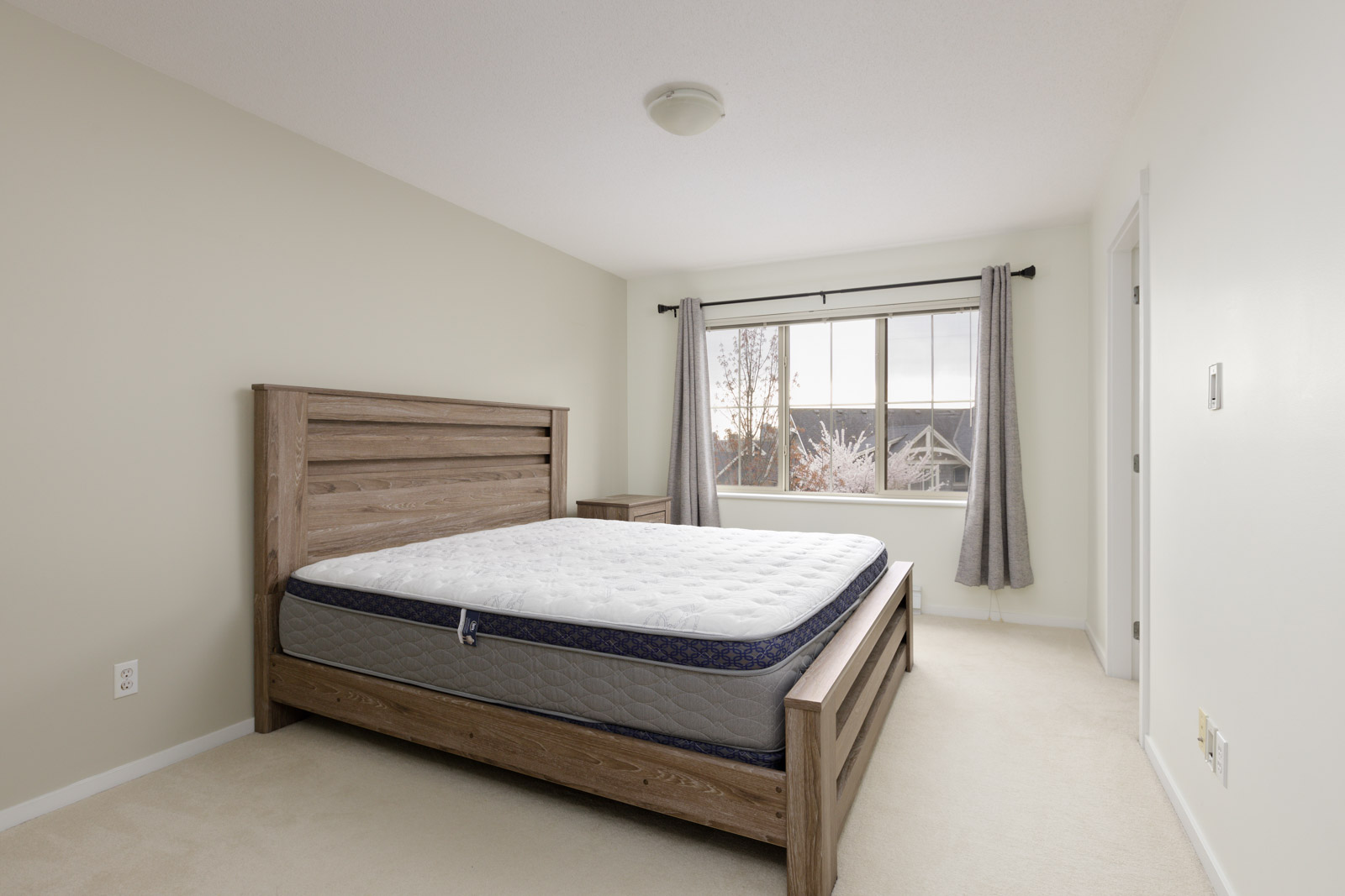 A bedroom with a wooden bed frame, a mattress without bedding, a nightstand, a window with gray curtains, and beige walls and carpet.