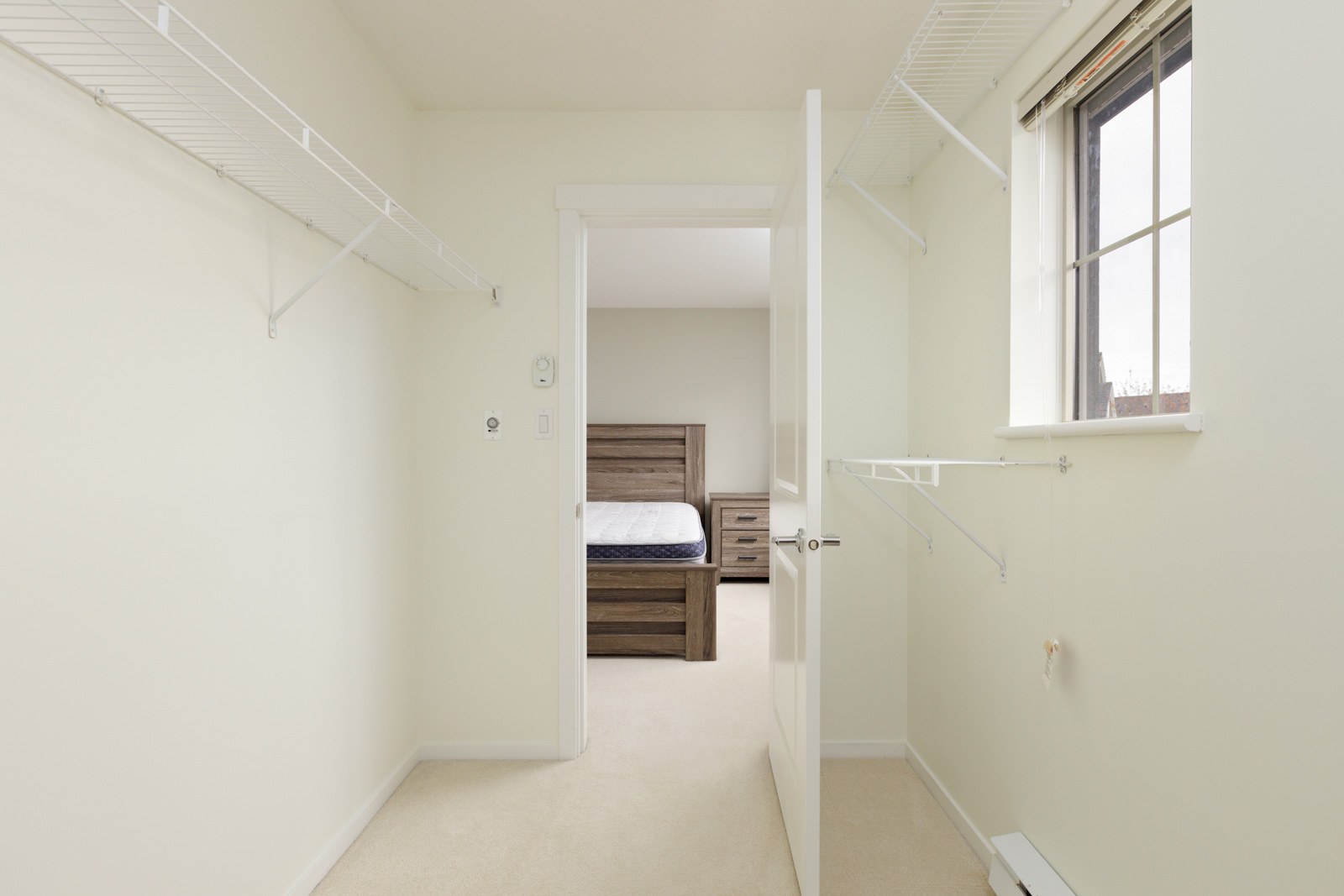 Empty walk-in closet with white wire shelves, open door, and view into bedroom with a wooden bed frame and nightstands.