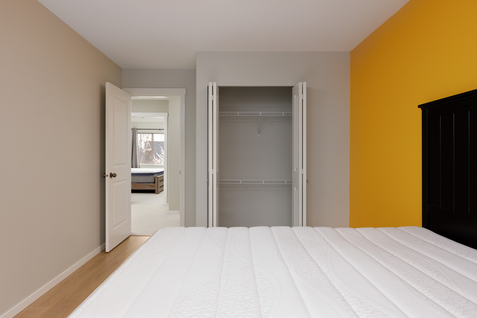 A bedroom with a bed, an open closet, a white door leading to another room with a bed, white walls, and a yellow accent wall.