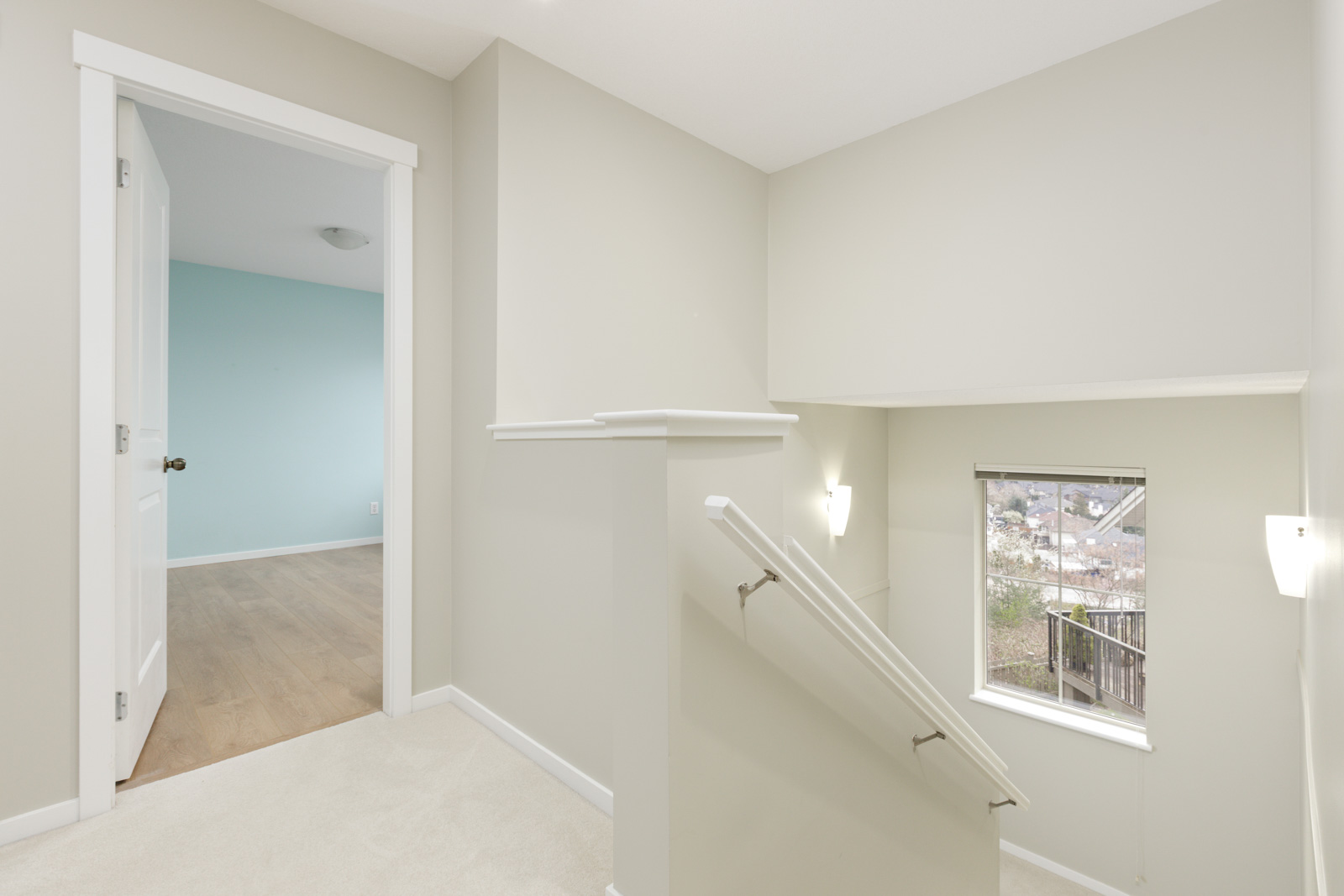 A carpeted staircase with a handrail leads to a landing area with a window and a door opening into a room with wooden floors and teal walls.